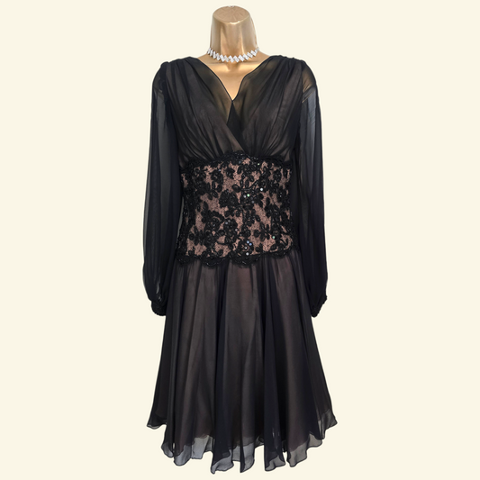 A black long-sleeve midi dress with a sheer mesh skirt, a deep V-neckline, and a fitted waist featuring black floral lace applique over a nude lining. The dress is displayed on a mannequin torso with a pearl necklace accessory, set against a solid cream background.