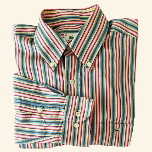 A folded long-sleeve striped dress shirt with pink, blue, yellow, and green vertical stripes, featuring cream buttons and a small green crocodile logo on the cuff, set against a solid cream background.