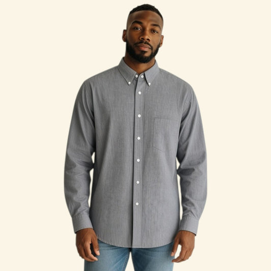 A man wearing a light gray long-sleeve button-down shirt with white buttons and blue jeans, standing against a solid cream-colored background.