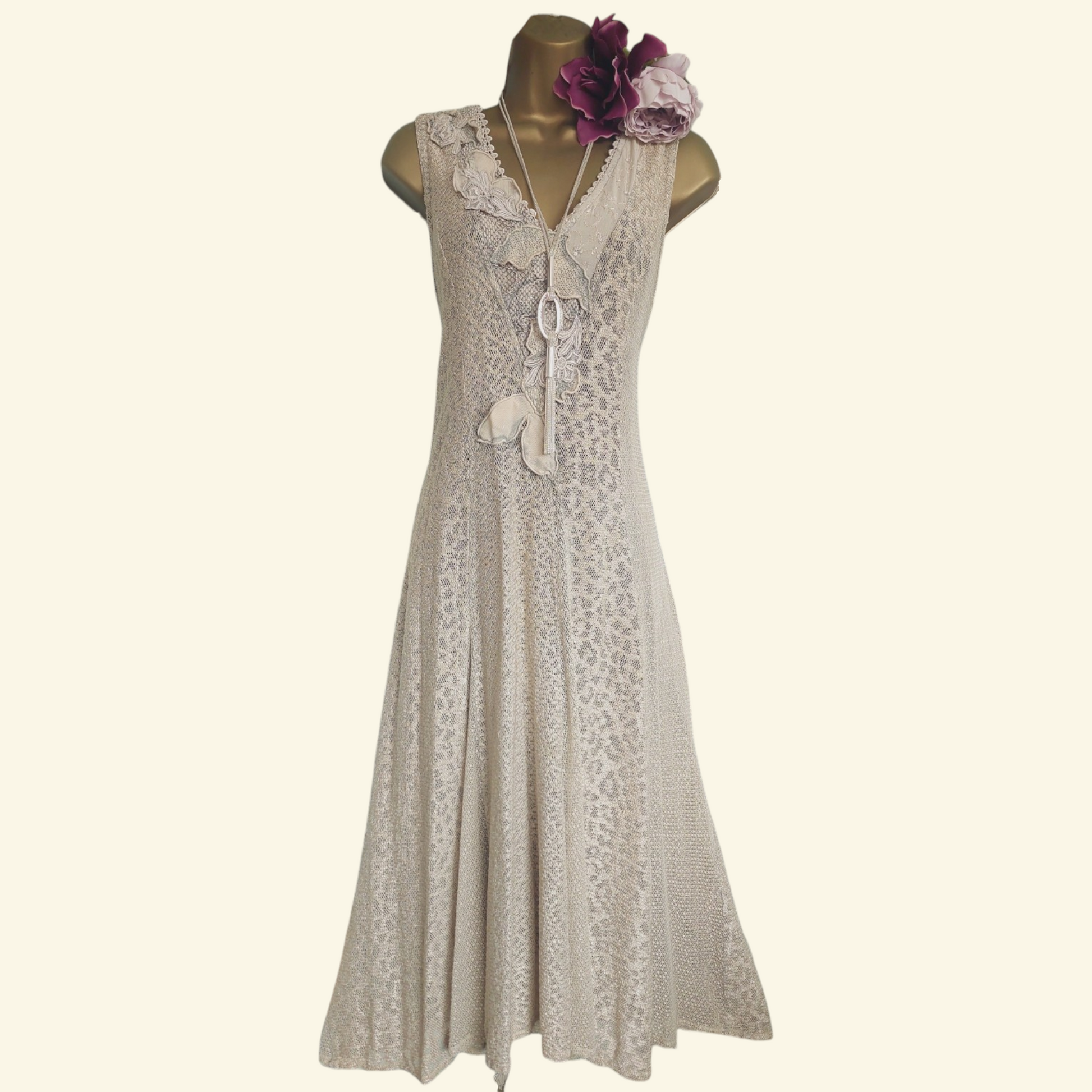 A sleeveless cream lace dress displayed on a mannequin with a gold neck collar and a purple flower accessory, set against a solid cream background.