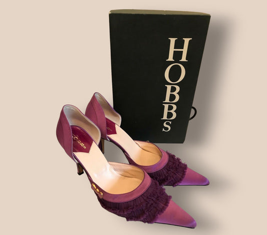 Purple high-heeled shoes with ruffles in front of a black 'Hobbs' box on a beige background