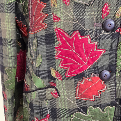 Close-up of a jacket featuring a green and black plaid base with vibrant red maple leaves and green oak leaves, accented by dark gray buttons.