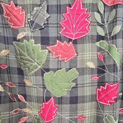 Close-up of a jacket featuring a green and black plaid base with vibrant red maple leaves and green oak leaves. 