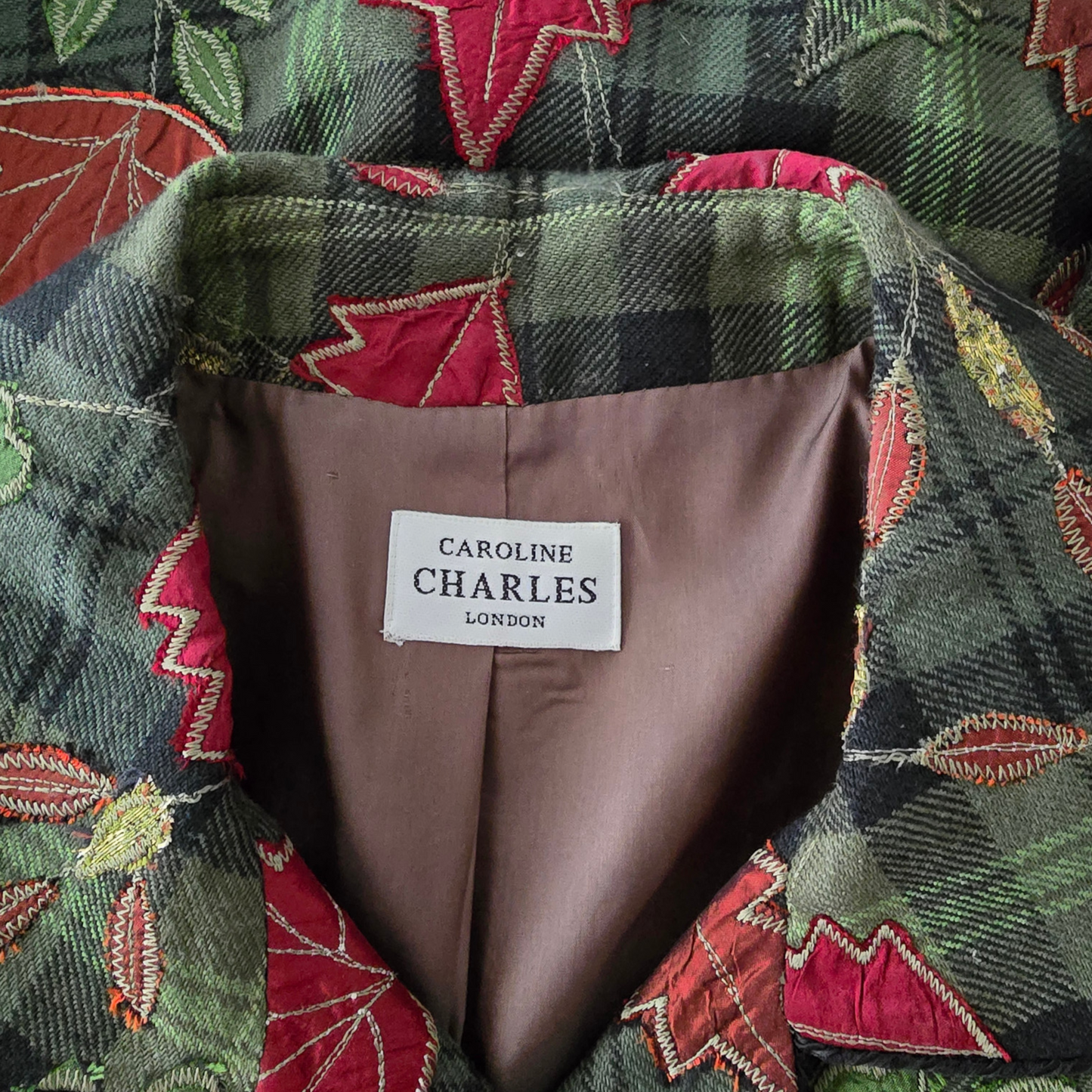 Close-up of a  jacket collar showing a white label that reads 'CAROLINE CHARLES LONDON' on a brown lining, with a green and black plaid and red floral quilted pattern visible around the collar.