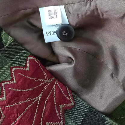 Close-up of a garment interior showing a taupe satin-like lining, a white size tag reading UK 12 EUR 38 USA 8, a black care label, and part of a red embroidered leaf motif over a green plaid fabric.