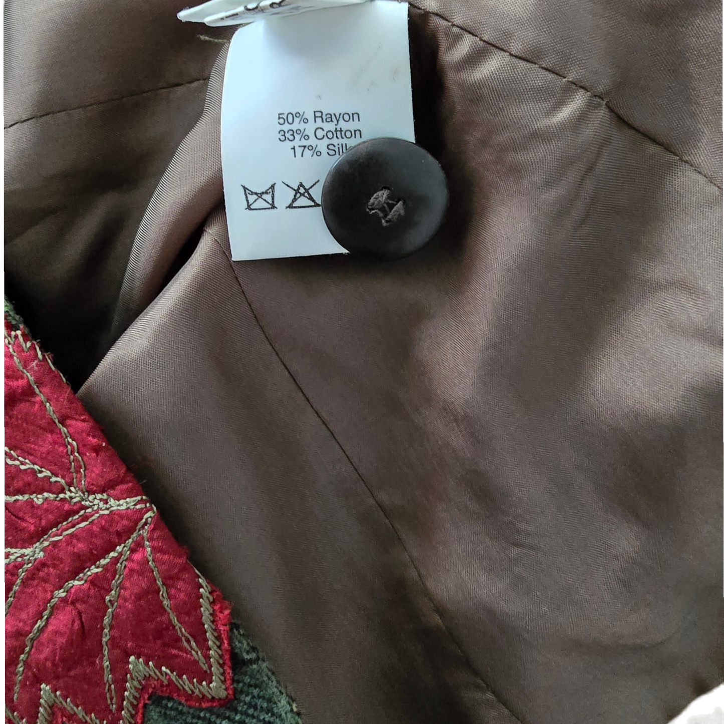 Close-up of a garment's interior label and button, showing a brown satin-like fabric with a white paper tag reading 50% Rayon, 33% Cotton, 17% Silk, and a dark brown button.