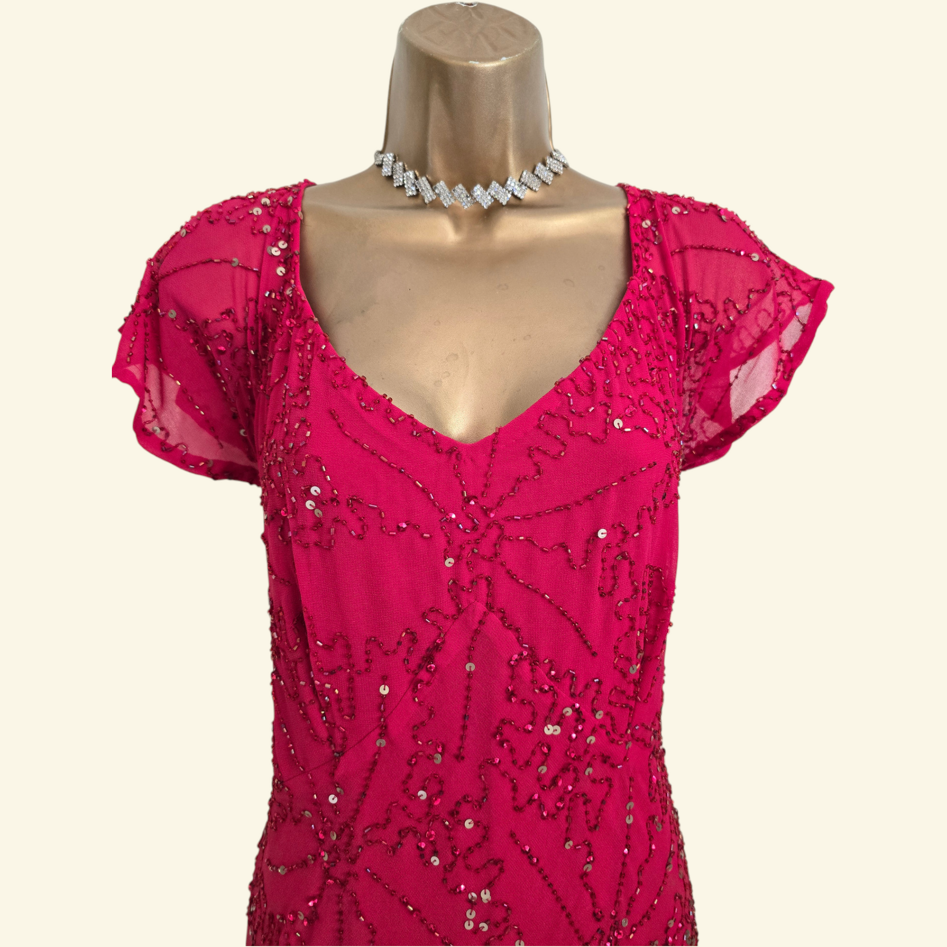 A bright pink beaded dress with flutter sleeves and a V-neckline, displayed on a gold mannequin torso with a silver rhinestone choker necklace against a light cream background.