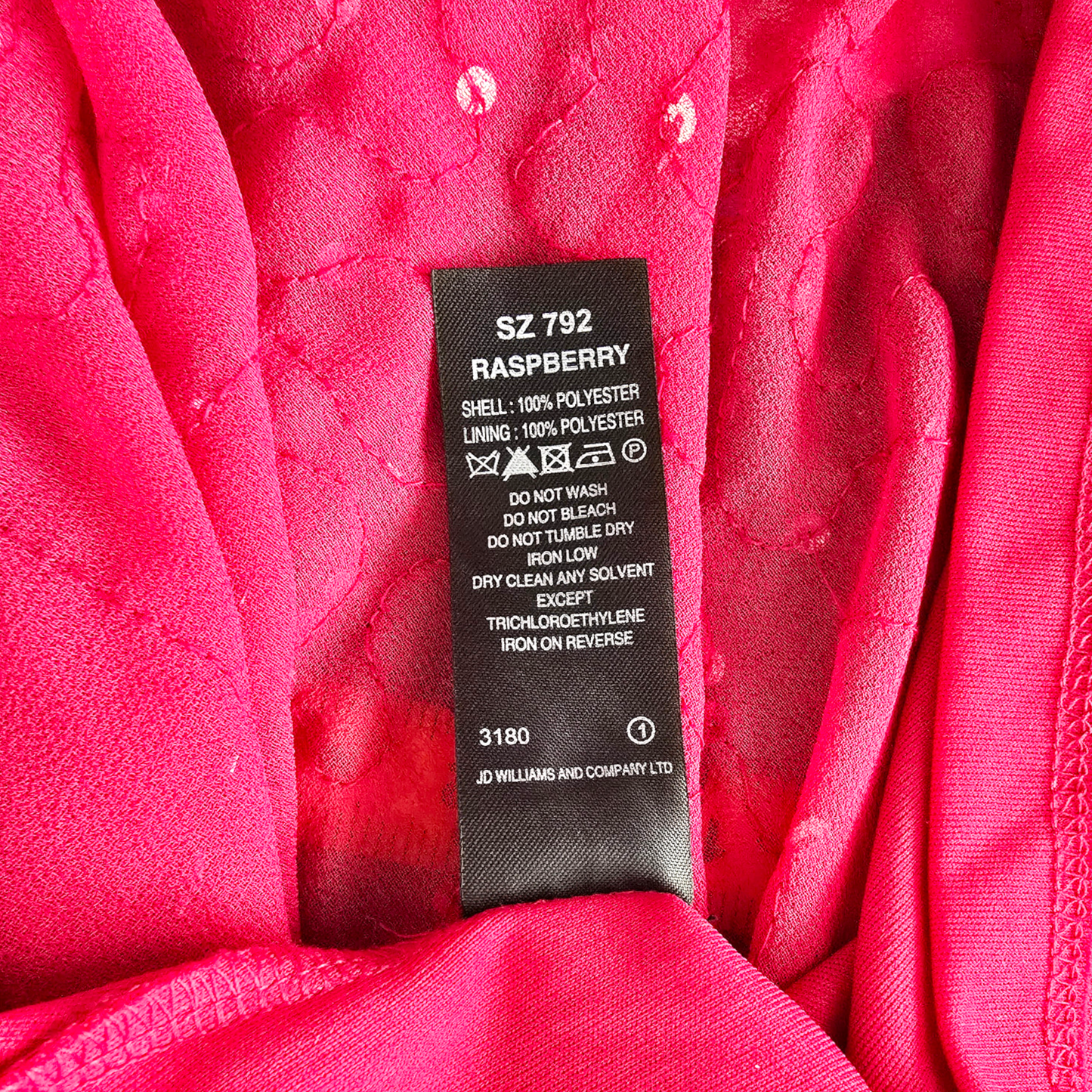 Close-up of a bright pink garment interior with a black sewn-in size and care label reading SZ 792 RASPBERRY, plus fabric texture and stitching visible.