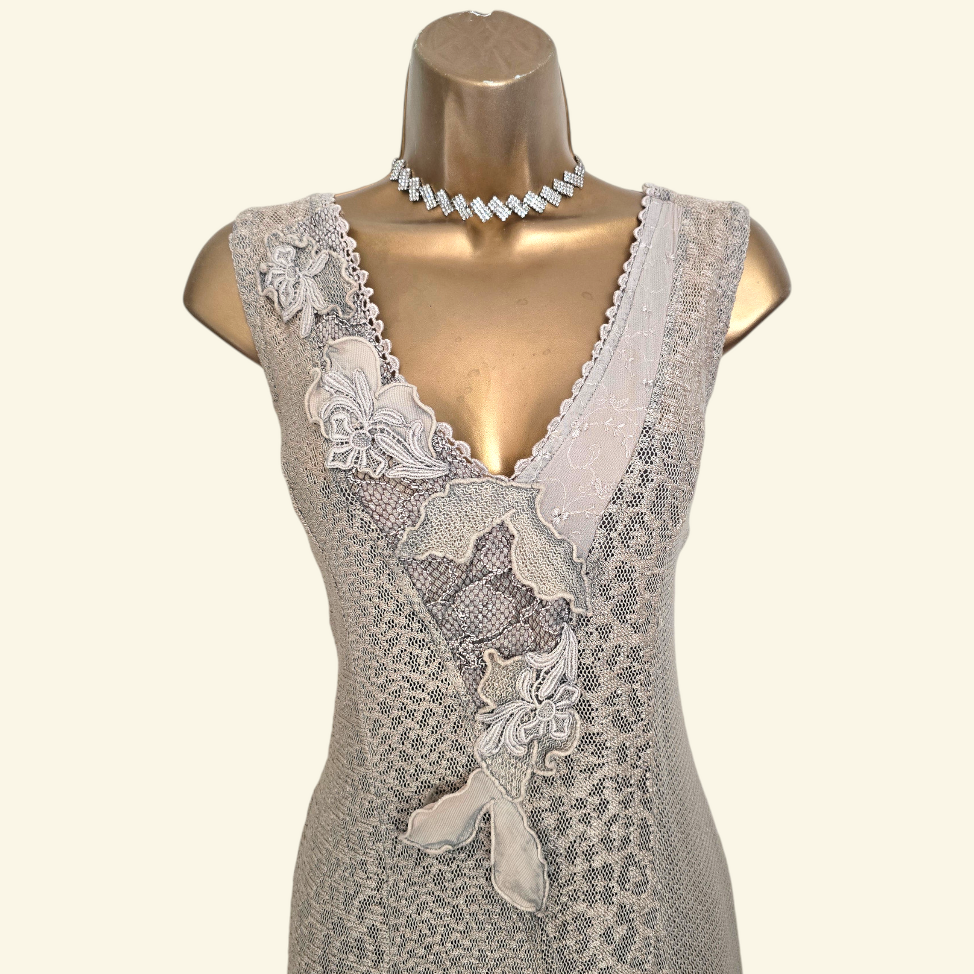 A sleeveless lace dress displayed on a metallic gold mannequin torso, featuring a deep V-neckline, floral applique details, and a textured pattern, paired with a silver rhinestone choker necklace.