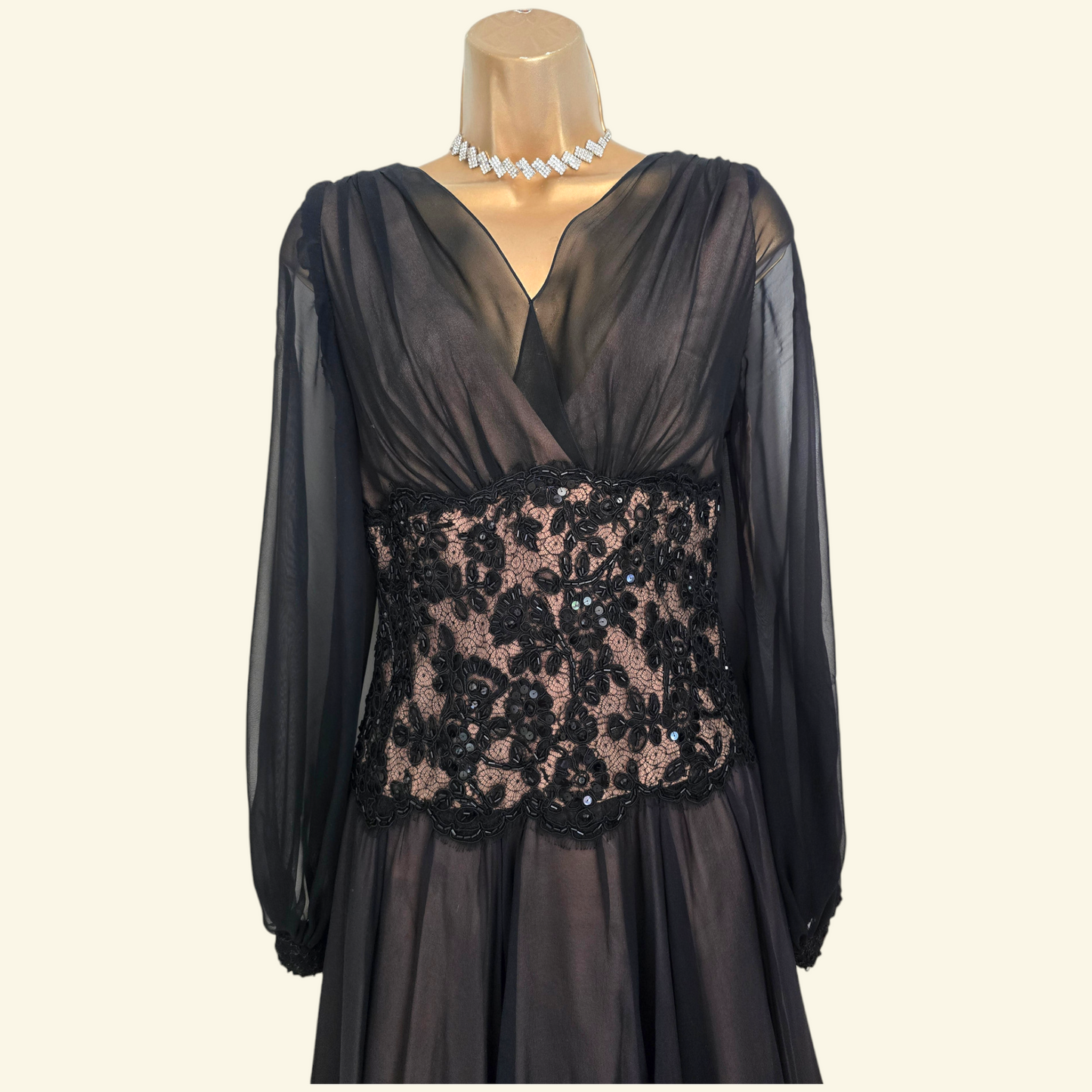 A black sheer long-sleeve evening dress displayed on a mannequin torso with a pearl necklace. The dress features a V-neckline, a fitted waist, and a prominent black floral lace panel with sequins across the bodice, set against a solid light beige background.