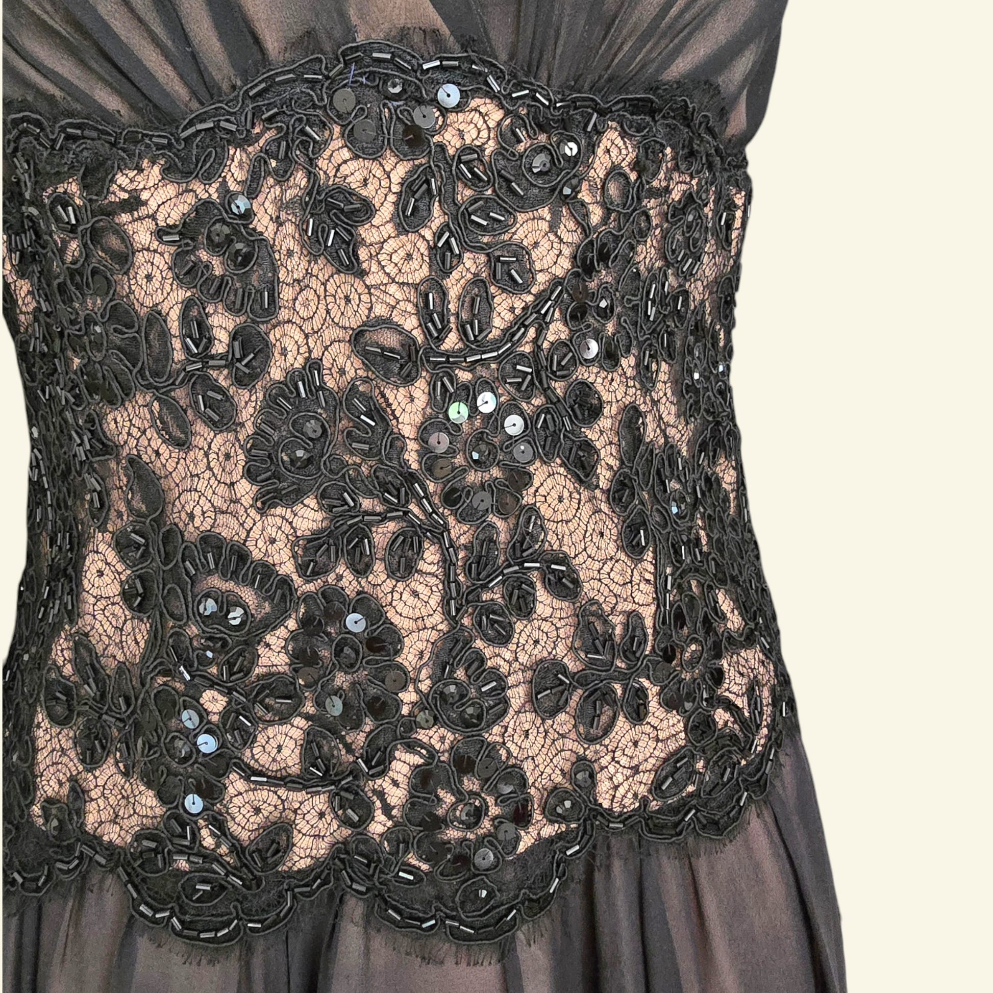 Close-up of a black lace bodice with floral embroidery and sequined details over sheer nude-toned underlay, shown on a dress against a light cream background.