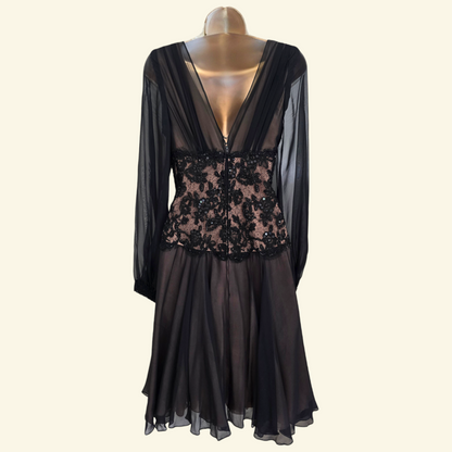 A black sheer long-sleeve dress displayed on a mannequin against a light beige background. The dress features a deep V-back, a sheer mesh overlay, and a fitted bodice with black floral lace applique over a nude lining.