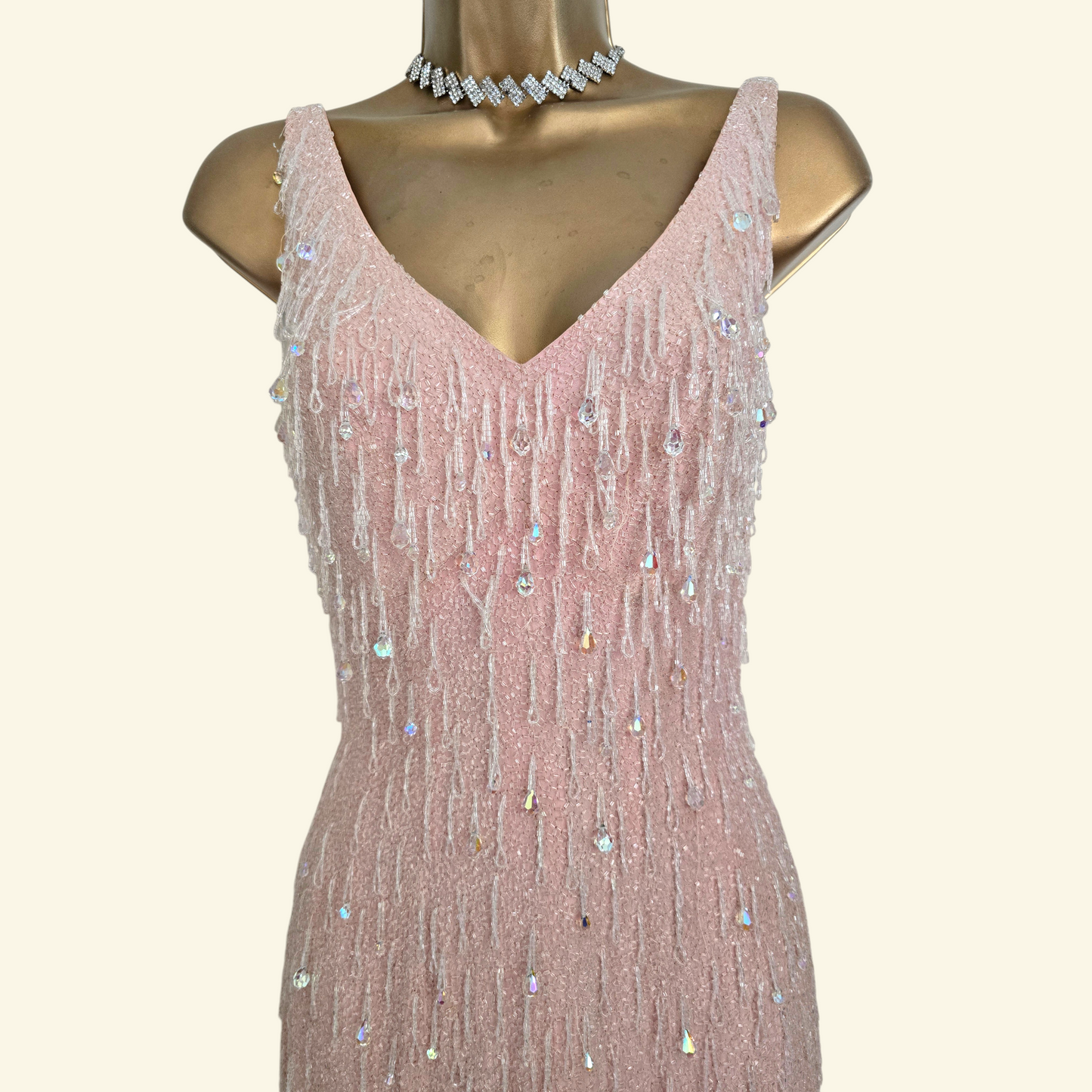 A sleeveless pink evening gown with a V-neckline and cascading beaded fringe embellished with rhinestones, displayed on a gold mannequin torso against a light beige background.