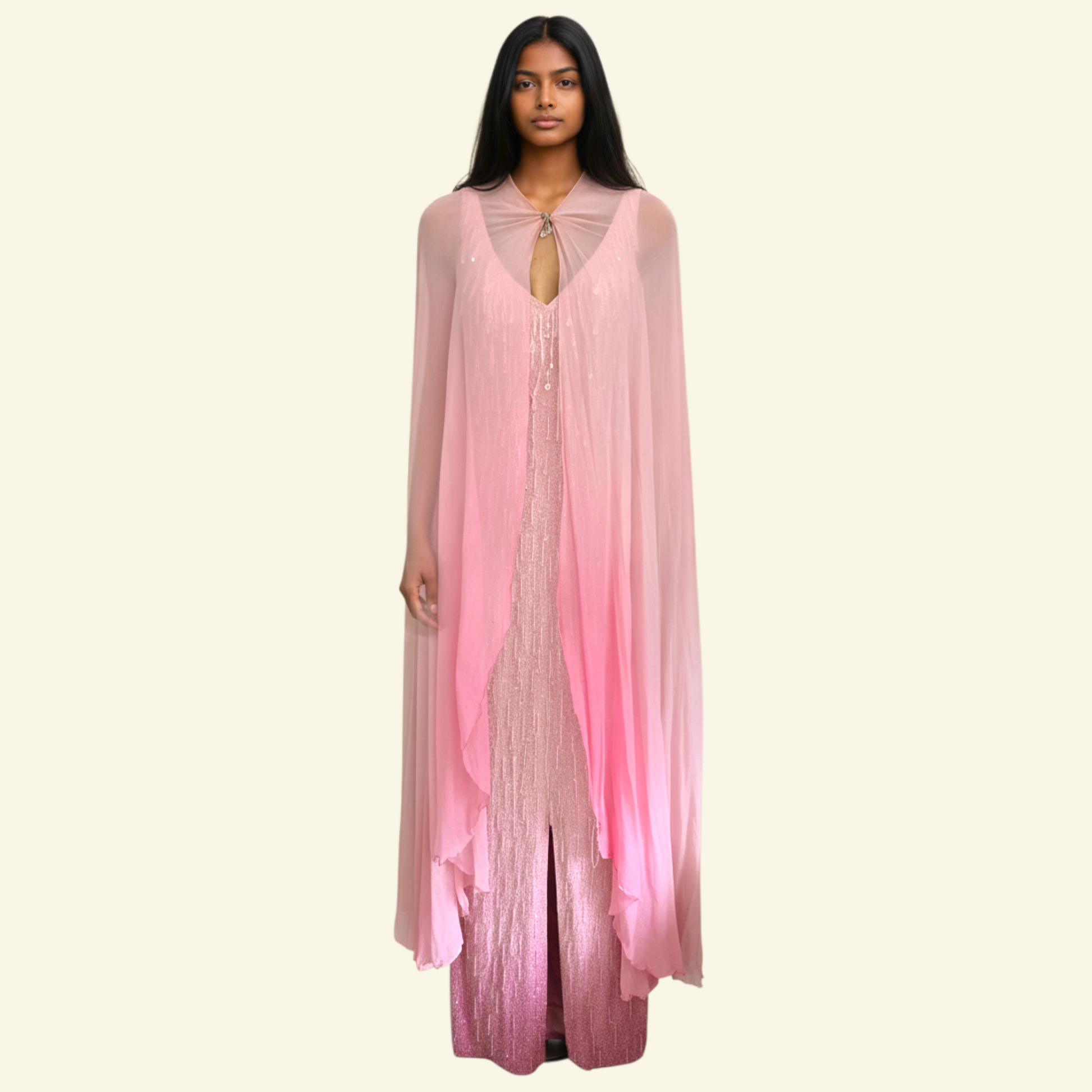 A woman modeling a long, flowing pink garment with a sheer draped overlay and a beaded inner layer, set against a solid cream background.