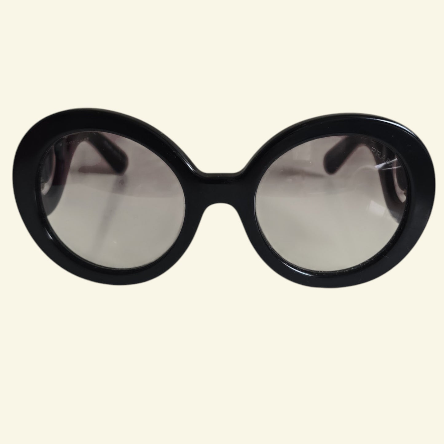 Oversized round sunglasses with a thick black frame and light gray tinted lenses, shown against a solid cream background.