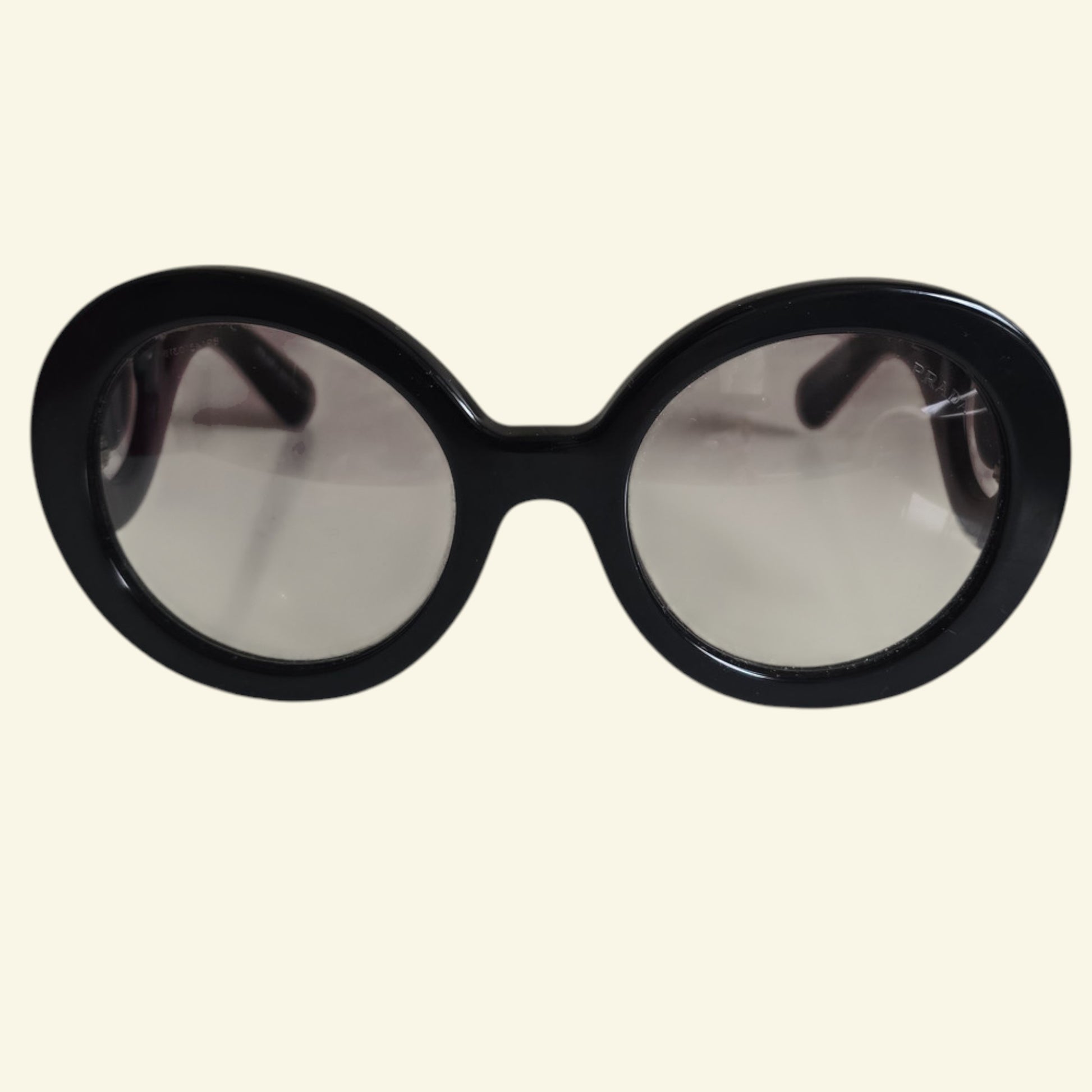 Oversized round sunglasses with a thick black frame and light gray tinted lenses, shown against a solid cream background.