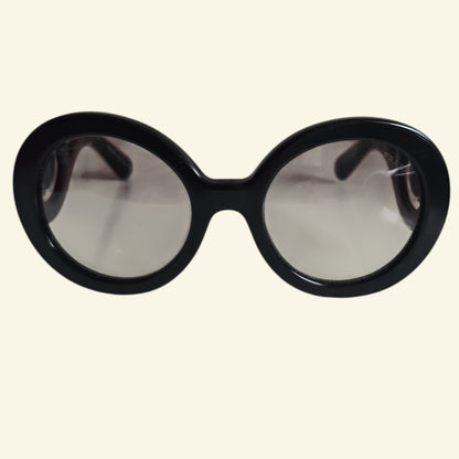 Oversized round sunglasses with a thick black frame and light gray tinted lenses, shown against a solid cream background.