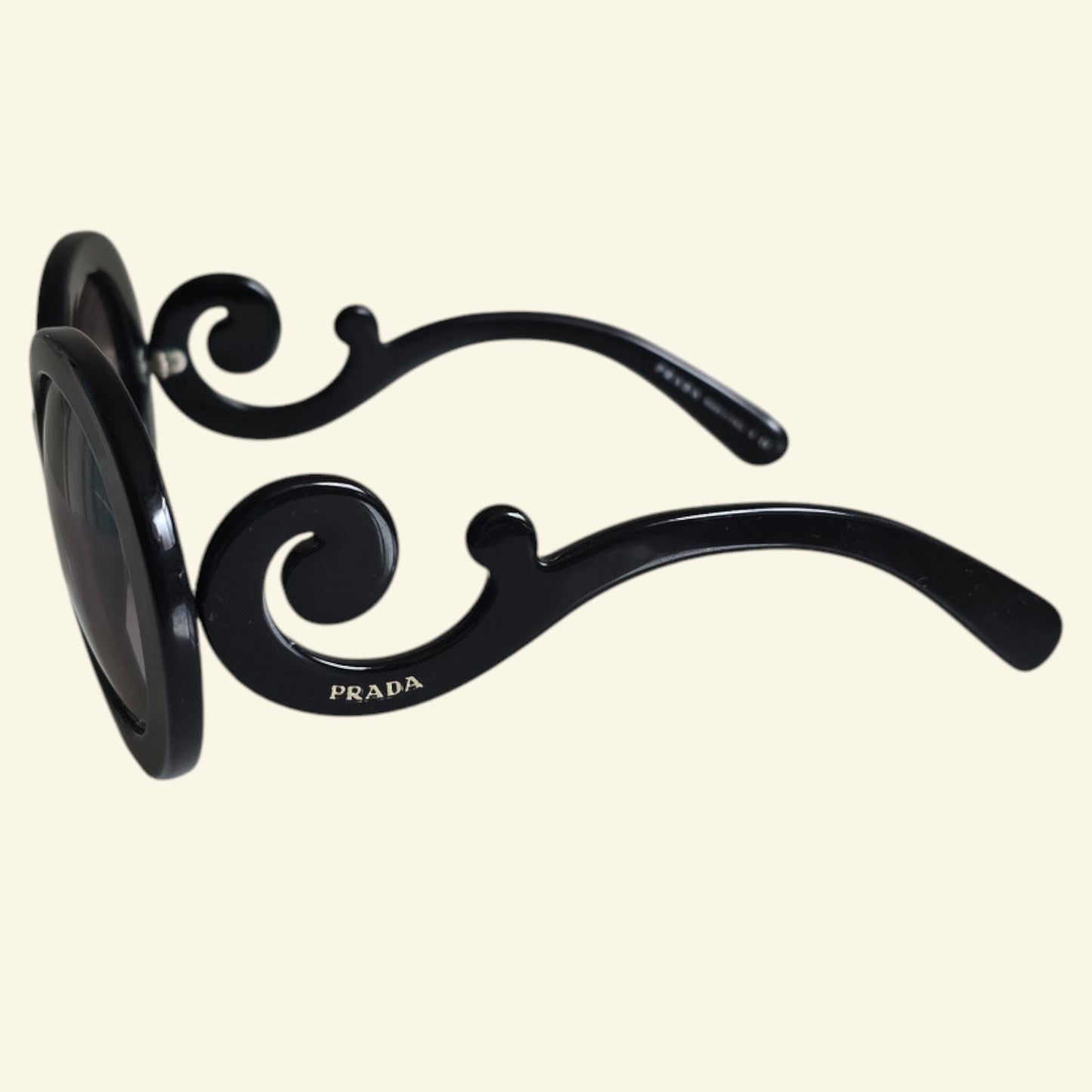Black Prada sunglasses with an ornate scrollwork design on the temples, shown against a solid cream background.