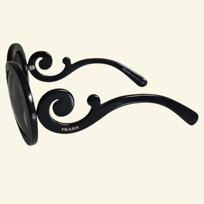 Black Prada sunglasses with an ornate scrollwork design on the temples, shown against a solid cream background.