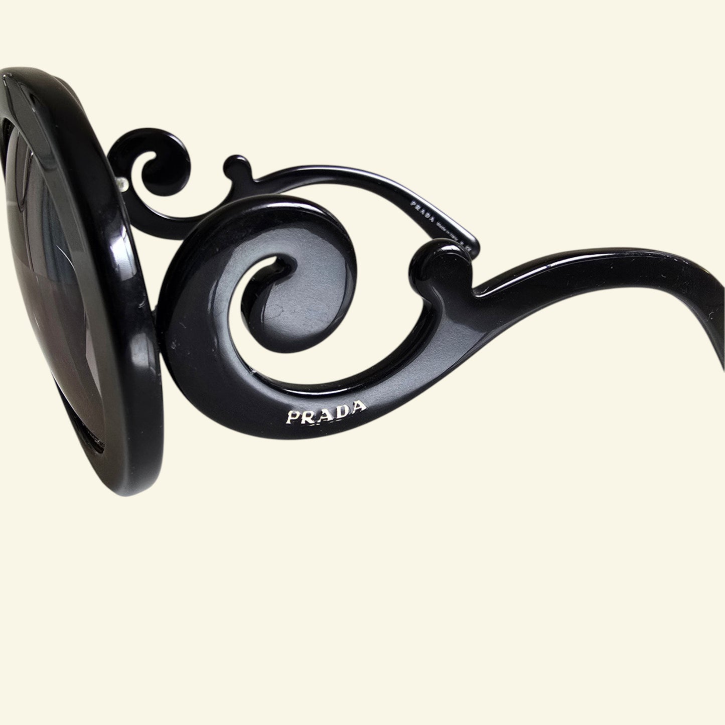 Close-up of black Prada sunglasses with an ornate scrollwork frame design and the PRADA logo printed on the temple, set against a solid cream background.