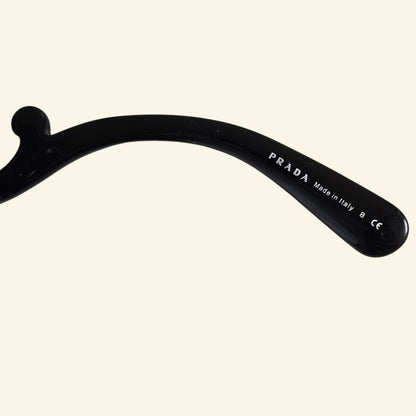 Close-up of a black Prada eyeglass arm with white printed branding and text, shown against a plain off-white background.