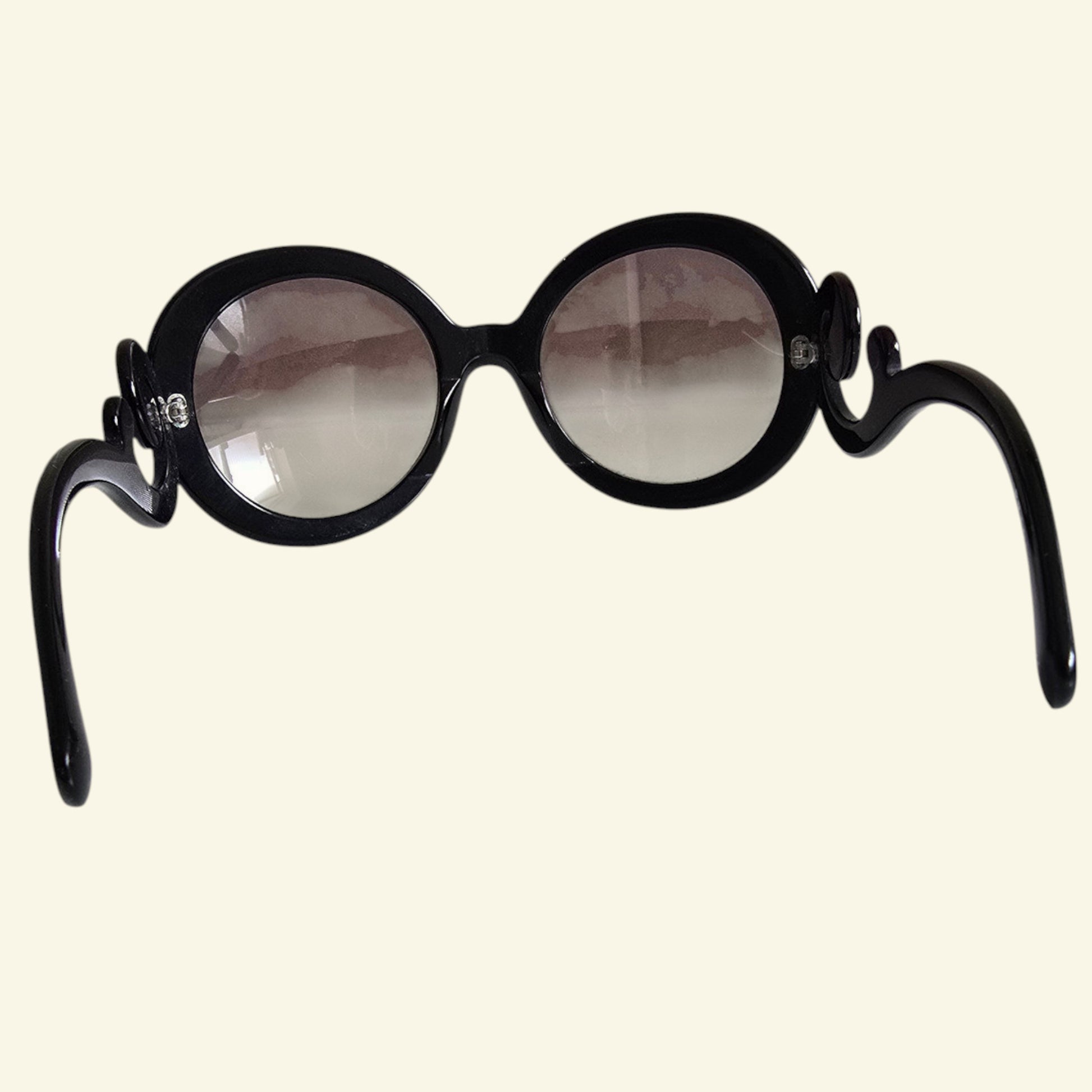 Black round sunglasses with decorative scroll details on the temples, shown against a solid cream background.