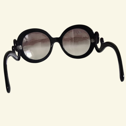 Black round sunglasses with decorative scroll details on the temples, shown against a solid cream background.