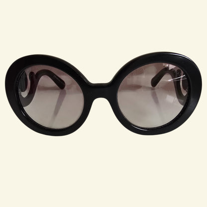 Oversized round sunglasses with a thick black frame and gradient tinted lenses, shown against a solid cream background.