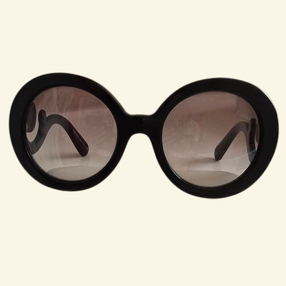 Oversized round sunglasses with a thick black frame and gradient brown lenses, shown against a solid cream background.