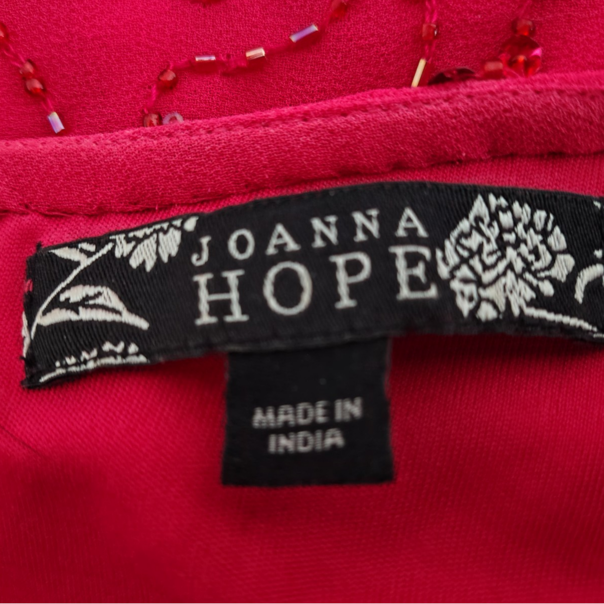 Close-up of a bright pink garment label reading JOANNA HOPE and MADE IN INDIA, with floral embroidery on the black tag and red beaded trim visible above.