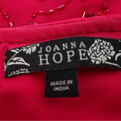 Close-up of a bright pink garment label reading JOANNA HOPE and MADE IN INDIA, with floral embroidery on the black tag and red beaded trim visible above.