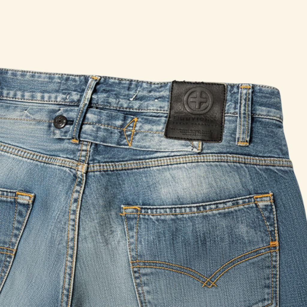 Close-up of the back waistband and pockets of light blue denim jeans, showing a black leather patch with a circular cross logo on the waistband.