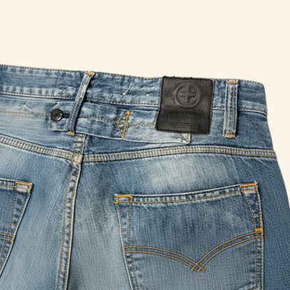 Close-up of the back waistband and pockets of light blue denim jeans, showing a black leather patch with a circular cross logo on the waistband.