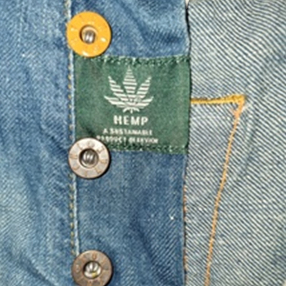 Close-up of a blue denim garment showing a green fabric label with a white hemp leaf and the text 'HEMP A SUSTAINABLE PRODUCT REVISION', plus brass and silver buttons.