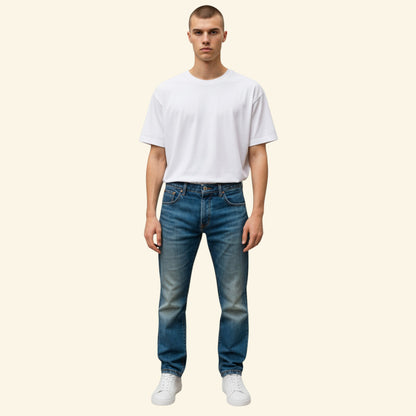 A man modeling a white crewneck t-shirt and blue jeans against a solid cream background.